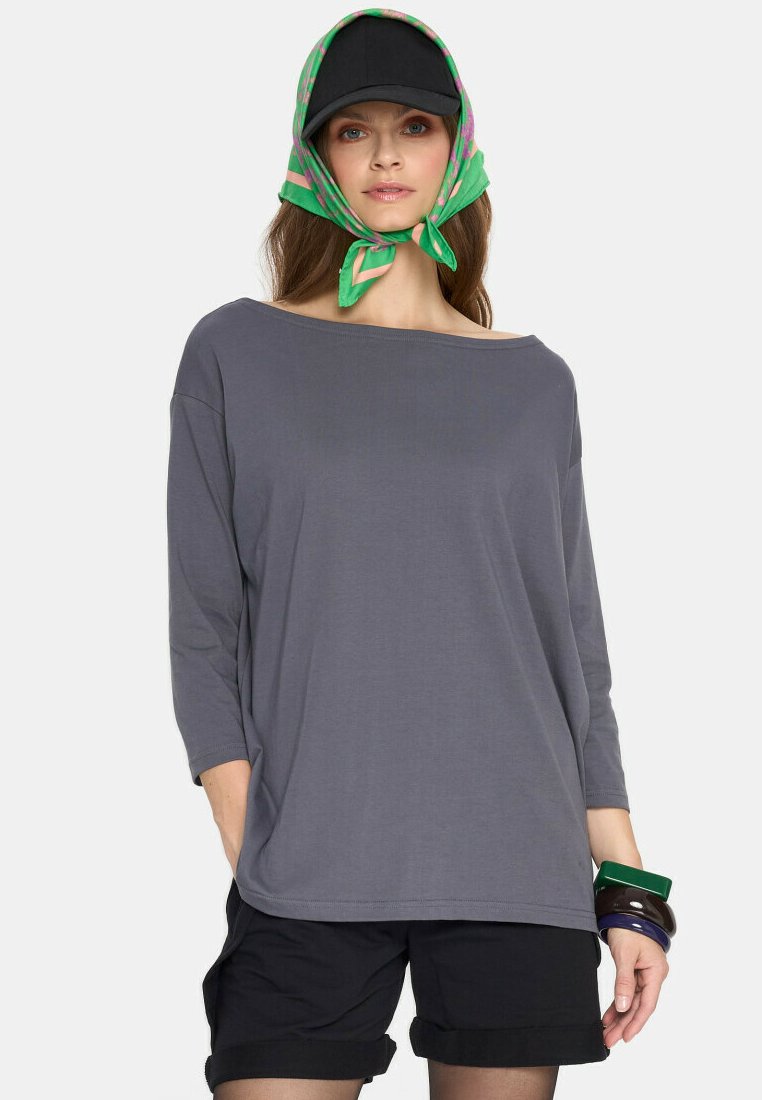 Szara, luźna bluzka z długim rękawem i szerokim dekoltem, stylizowana z czarnymi szortami. Dodatki to czapka w kolorze czarnym oraz zielony szalik w wzory.