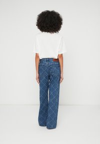 Calças de ganga azul boca de sino com padrão em diamante, com um remendo de couro castanho na cintura e dois bolsos traseiros. Combinadas com uma t-shirt branca.