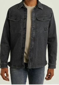 Camicia di jeans nera con due tasche sul petto, chiusura con bottoni e texture sbiadita. Presenta un colletto a punta e cuciture a contrasto. Indossata sopra un top chiaro.