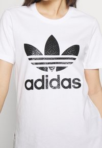 Biała koszulka z okrągłym dekoltem z teksturowanym czarnym logo Adidas z motywem liścia i trzema poziomymi paskami poniżej logo.