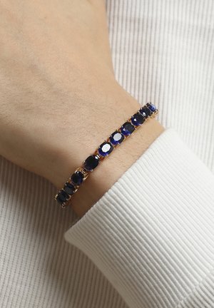 Bracciale in tono oro con pietre blu ovali e rotonde, montate in impostazioni a baffo, indossato su un polso con una manica in tessuto bianco testurizzato.