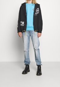 Světle modrý svetr nošený pod černou bundou s grafickými potisky, doplněný o vybledlé modré džíny a černé chelsea boty.