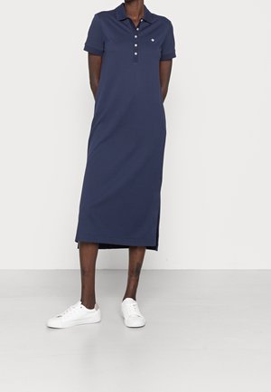 Person trägt ein marineblaues Polo-Midikleid mit kurzen Ärmeln und weißen Sneakers, steht vor einem schlichten Hintergrund.