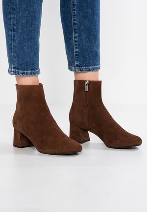 Braune Wildleder-Stiefeletten mit Blockabsätzen und seitlichen Reißverschlüssen, getragen zu verkürzten blauen Jeans auf weißem Hintergrund.