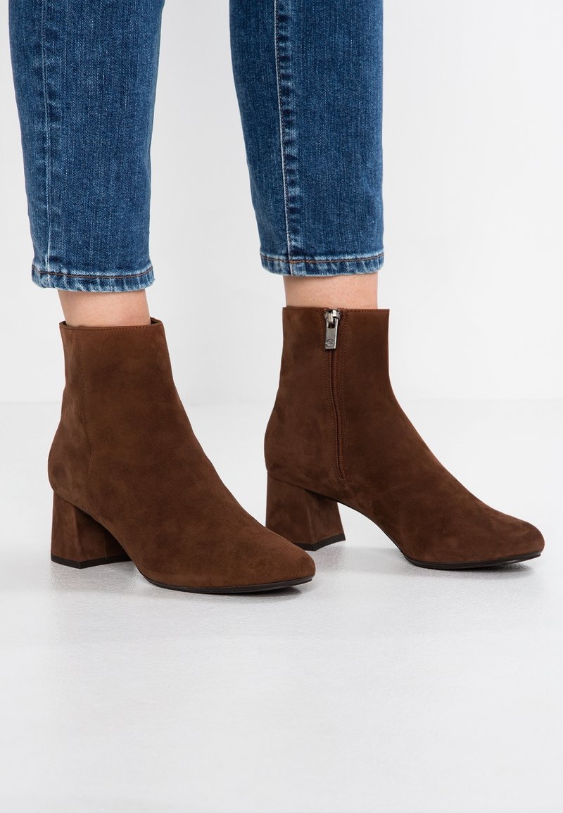 Bottines en daim marron avec talons carrés et fermetures éclair latérales portées avec un jean bleu court sur fond blanc.