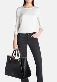 Borsa a mano in pelle nera con hardware dorato e doppi manici, con una texture liscia e una fodera viola, tenuta da un modello che indossa una maglietta bianca.