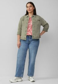 Olivgrüne Denimjacke über einem pinken gemusterten Top, kombiniert mit hellblauen Weitbeinjeans und grauen Sneakers mit weißen Akzenten.