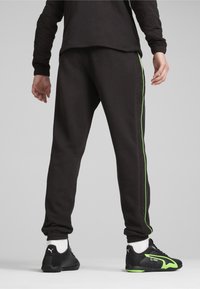 Zwarte trainingsbroek met een slanke pasvorm, voorzien van een groene zijstreep en textuuraccenten langs de buitenste naden. Gecombineerd met zwarte sportschoenen.