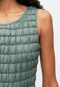 Artimas vaizdas į asmenį, vilkintį be rankovių, tekstūruotą šviesiai žalią marškinėlį su surinktomis, pūkuotomis horizontaliomis eilėmis ir matomais petimi ir viršutine ranka.