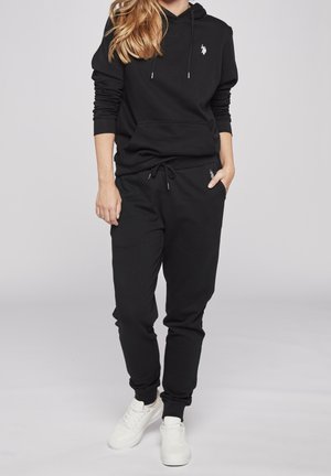 Hoodie noir avec une poche avant et un cordon de serrage, associé à un jogger noir assorti avec une taille élastique et un accent logo. Baskets blanches.