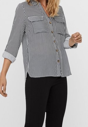 Femme portant une chemise à rayures verticales noires et blanches boutonnée, manches retroussées, et un pantalon noir ajusté sur un fond uni.