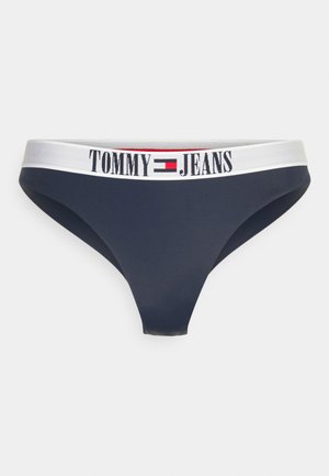 Marinblå tanga med en bred vit midjeband där "TOMMY JEANS" är tryckt med svart. Tillverkad av mjukt, elastiskt tyg.