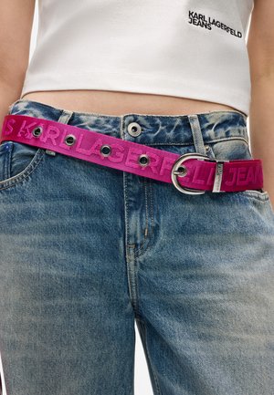 Różowy pasek z materiału z wytłoczonym napisem "KARL LAGERFELD JEANS", z metalową klamrą w kolorze srebrnym i okrągłym elementem, noszony na niebieskich dżinsach.