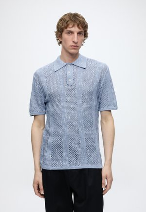 Jeune homme portant un polo en maille bleu clair à manches courtes avec un motif en maille ouverte texturée et un pantalon noir, debout devant un fond uni.