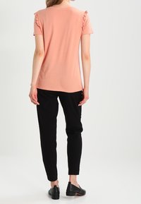 Hlava s krátkým rukávem z broskvového materiálu s volánkovými detailními rameny, nošená s černými kalhotami a plochými černými botami. Jednoduchý design, jemná textura.