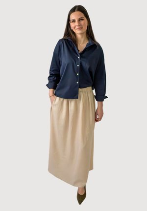 Femme aux longs cheveux bruns portant une chemise à boutons bleu marine, une jupe beige longueur cheville et des chaussures pointues vert olive, debout devant un fond uni.