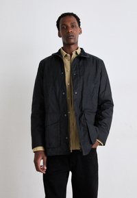 Veste noire imperméable au design légèrement structuré, avec des boutons-pression, deux poches avant et un col, portée par-dessus une chemise olive.