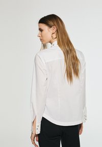 Camicia bianca a maniche lunghe con colletto alto, dettagli a fessura sulle maniche e accenti con bottoni dorati sui polsini, caratterizzata da una texture liscia.