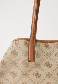 Beżowa torba na ramię z płótna z wytłoczonym wzorem logo, brązowymi skórzanymi akcentami i szwami. Posiada jeden, wydłużony pasek.
