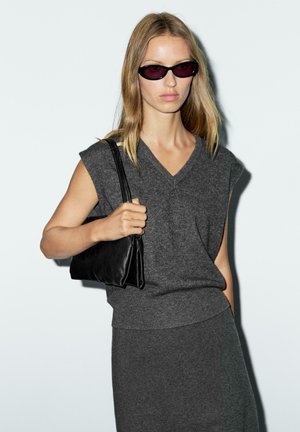 Woman with long blonde hair wearing dark sunglasses, sleeveless grey knit top, matching skirt, and holding a black shoulder bag.