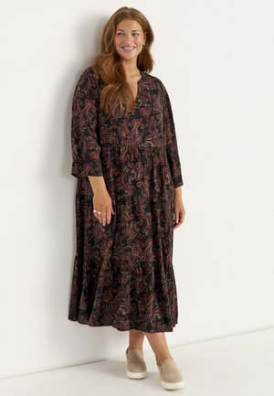 Kvinde med langt hår, der smiler, iført en mørk blomstret langærmet maxi-kjole og beige slip-on sko, står mod en hvid væg.