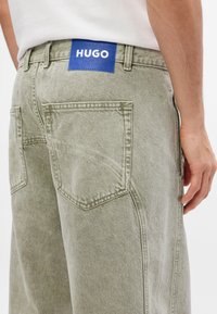Zaļas džinsa bikses ar teksturētu apdari, kurām ir divi aizmugurējie kabatas un zils etiķete ar uzrakstu "HUGO" baltā krāsā jostasvietā.