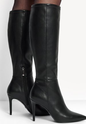 Bottes en cuir noir montant jusqu’aux genoux, à bouts pointus, talons aiguilles et fermetures éclair latérales, portées avec des bas foncés.