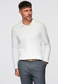 Witte lange mouwen polo met een platte kraag, van een gestructureerde stof, gedragen met grijze broek en een bruine riem, met ingewikkelde stikdetails.