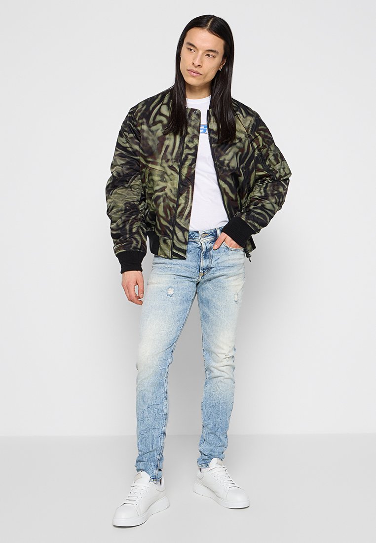 Giacca bomber camouflage in verde scuro e nero, t-shirt bianca, jeans chiari strappati, e sneaker bianche, in piedi davanti a uno sfondo neutro.