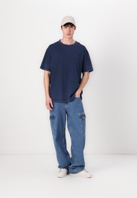 Námořnická modrá oversized tričko s malým logem, v kombinaci s světle modrými volnými cargo džíny s bočními kapsami a bílými teniskami.