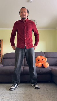 Camicia rossa con bottoni, jeans grigi e sneakers nere esposti su una parete verde chiaro, con un polpo di peluche arancione su un divano grigio.