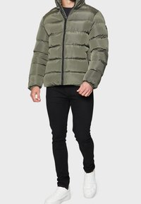 Oliwkowa kurtka puchowa z zamkiem z przodu, pikowany design i kaptur. Połączona z czarnymi spodniami typu slim fit i białymi trampkami.