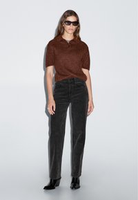 Lichtbruine gebreide polo met korte mouwen, gecombineerd met zwarte corduroy broek en zwarte enkellaarsjes met hak. Model staat tegen een grijze achtergrond.