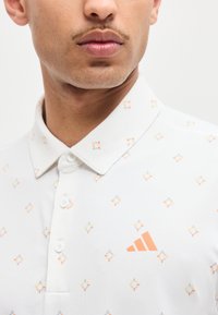 Άνδρας που φοράει λευκό πουκάμισο με γιακά, με μικρά πορτοκαλί και πράσινα σχέδια σταυρού και πορτοκαλί λογότυπο Adidas στο στήθος.