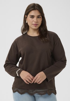 Ung kvinde med mørkebrunt langt hår iført en brun langærmet top med blonder og lyseblå jeans mod en ensfarvet baggrund.