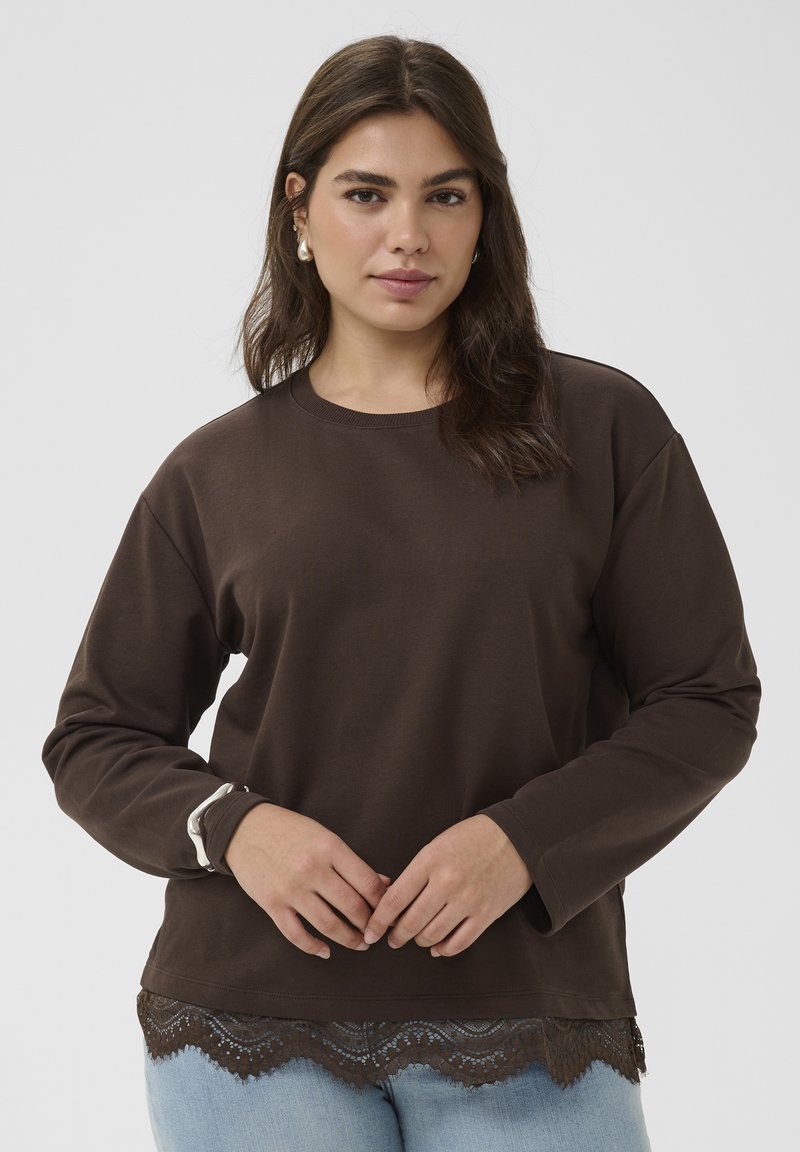 Jeune femme aux longs cheveux bruns foncés portant un haut marron à manches longues avec bordure en dentelle et un jean bleu clair, sur un fond uni.