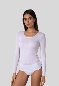 Donna con capelli scuri e lisci, che indossa un top bianco trasparente a maniche lunghe e biancheria intima bianca, in piedi davanti a uno sfondo grigio semplice.