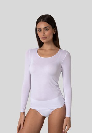 Donna con capelli scuri e lisci, che indossa un top bianco trasparente a maniche lunghe e biancheria intima bianca, in piedi davanti a uno sfondo grigio semplice.