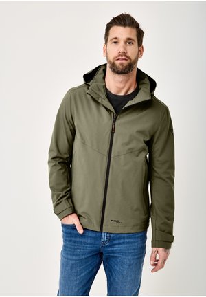 Man wearing an olive green hooded jacket with front zipper and blue jeans, standing against plain light background.