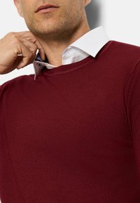 Burgunderpullover mit Rundhalsausschnitt, aus einem strukturierten Stoff, über einem frischen weißen Hemd mit blauem Akzent geschichtet.