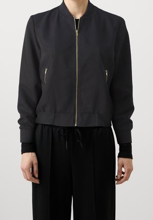 Vrouw draagt een zwart bomberjack met gouden rits en zijzakken met rits, gecombineerd met een zwarte broek met aantrekkoord in de taille.