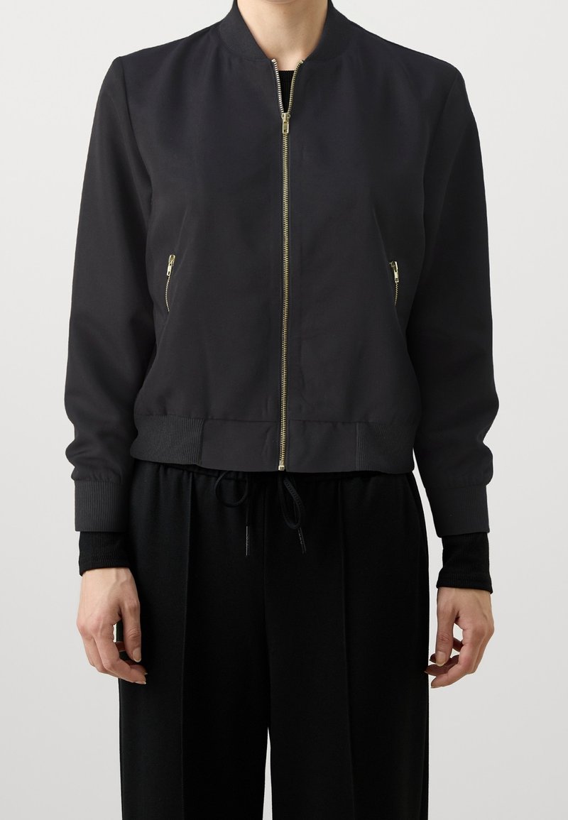 Femme portant une veste bomber noire avec une fermeture éclair dorée et des poches latérales zippées, associée à un pantalon noir avec taille à cordon.