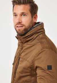 Brown jacket with a high collar, soft gray lining, zippered pockets, and a textured finish. Features a black logo patch on the sleeve.