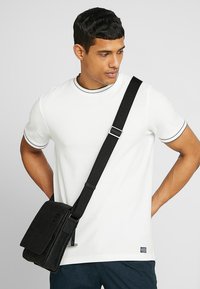 T-shirt bianco a maniche corte con bordi neri, abbinato a una borsa a tracolla nera con chiusura a pattina e tracolla regolabile.