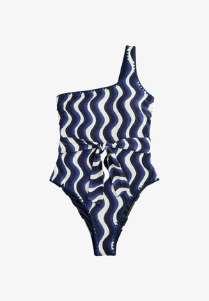 Frau trägt einen Badeanzug mit einem Träger und marineblauen und weißen wellenförmigen Streifen, von hinten vor einem unscharfen Außenhintergrund gezeigt.