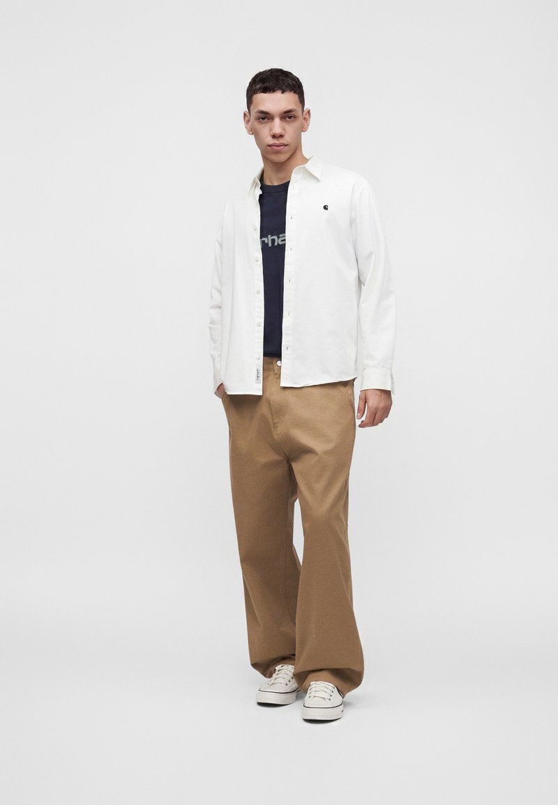 Jeune homme debout portant une chemise blanche boutonnée par-dessus un t-shirt noir, un pantalon marron ample et des baskets blanches sur un fond uni.