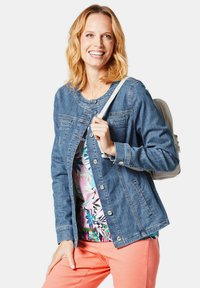 Jeansjacke mit Rundhalsausschnitt, silbernen Knöpfen und zwei Vordertaschen. Kombiniert mit einem blumigen Oberteil und korallenfarbenen Hosen. Backpack-Tragegurte sichtbar.