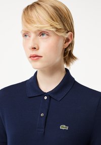 Granatowa koszulka polo z teksturowanej tkaniny, z zapięciem na trzy guziki i zielonym haftowanym logo krokodyla po lewej stronie klatki piersiowej.