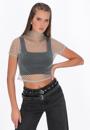 Junge Frau mit geflochtenem Haar, die ein durchsichtiges, gepunktetes Crop-Top über einem schwarzen Tanktop, schwarze mit Nieten besetzte Gürtel und schwarze Hosen trägt, posiert mit einer Hand in der Hüfte.