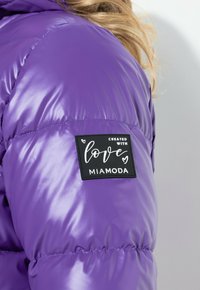 Chaqueta acolchada morada con textura brillante, que presenta una etiqueta negra con la frase "Creada con amor" y el nombre de la marca "Miamoda" en la manga.